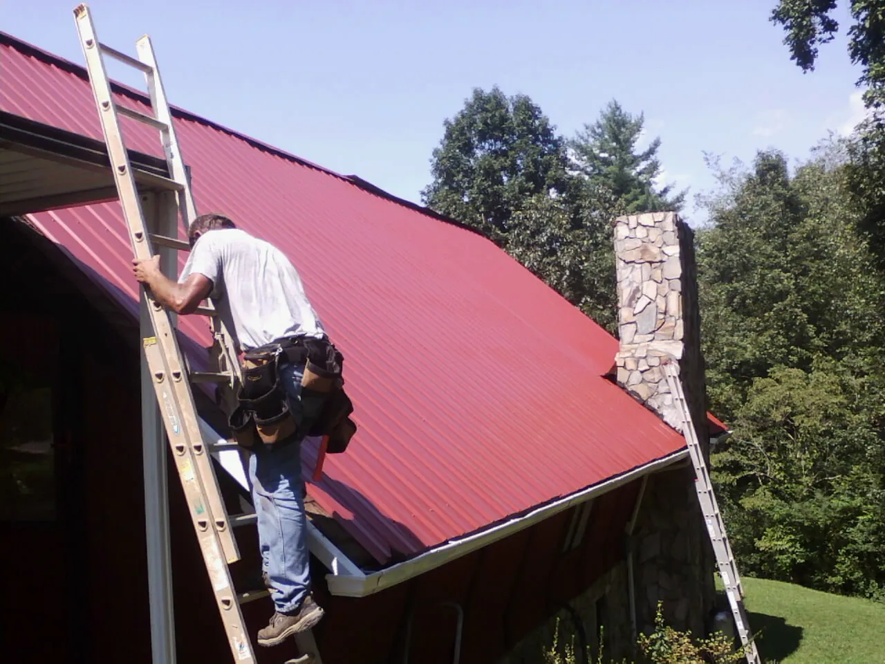 Roofer on a ladder beside a red metal roof and stone chimney — Soffit & Fascia Repair by Roofing Brothers in West Nyack